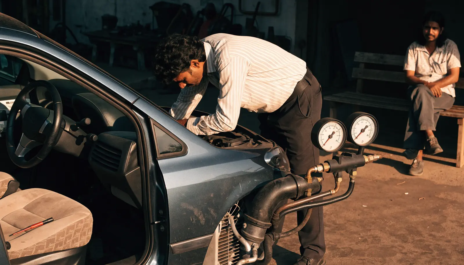 A technician checks a hatchback’s A/C with gauges connected and a thermometer on the seat while the owner waits in the shade.