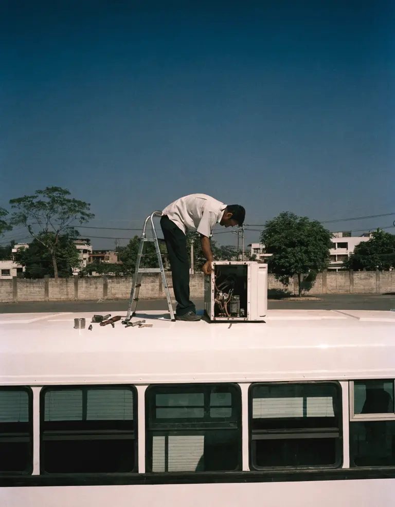 A technician on a ladder working at the roof-mounted A/C unit of a plain white bus in a yard.
