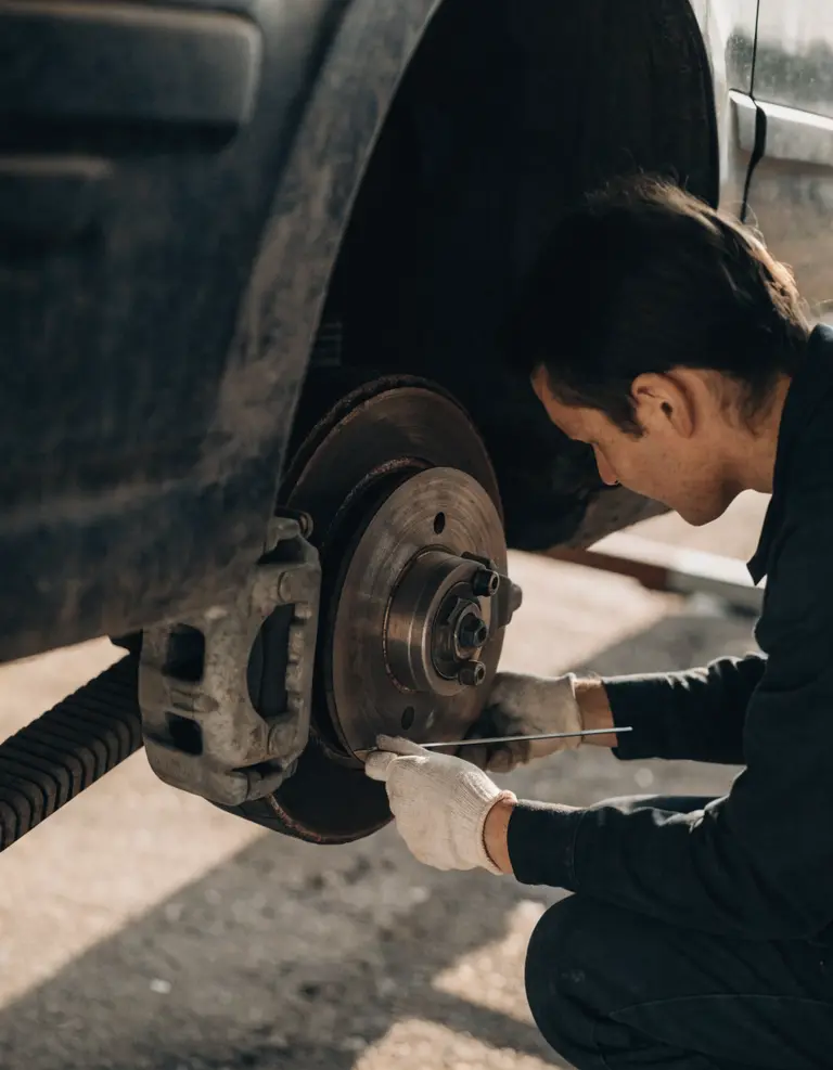 A technician beside a raised car with the wheel removed, measuring brake pad thickness against the disc.
